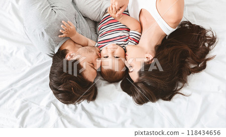 A happy family of three is lying in bed together. The parents are kissing their newborn baby on the cheeks. The baby is wearing a striped shirt and is looking up at the camera 118434656