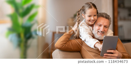 A grandfather and his granddaughter enjoy a happy moment together, laughing as they explore a tablet. The cozy living room, with soft lighting, creates a warm atmosphere. A grandfather and his granddaughter enjoy a happy moment together, laughing as they explore a tablet. The cozy living room, with soft lighting, creates a warm atmosphere. 118434673