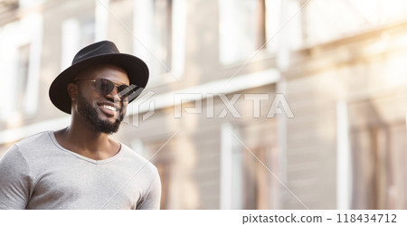 A man wearing a hat and sunglasses walks down a city street. He is smiling and looking off to the side. A man wearing a hat and sunglasses walks down a city street. He is smiling and looking off to the side. 118434712