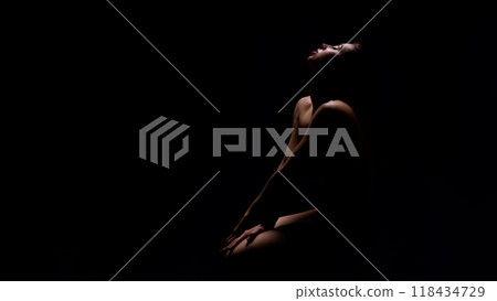 Naked young woman sitting in darkness with her arms on knee and looking up, panorama with copy space 118434729