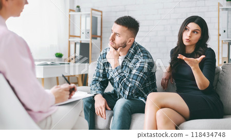 A couple sits on a couch in an office, talking to a therapist. The woman is talking and gesturing with her hand, while the man is listening and thinking thoughtfully. The therapist is taking notes. 118434748
