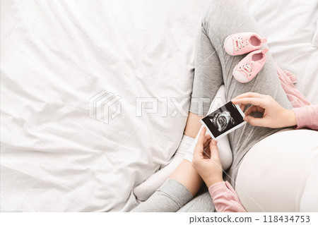 A pregnant woman lounges on a bed in the morning, gently holding an ultrasound picture of her baby, with baby shoes nearby, view above, copy space A pregnant woman lounges on a bed in the morning, gently holding an ultrasound picture of her baby, with baby shoes nearby, view above, copy space 118434753