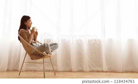 A woman sits in a chair by a window, enjoying a warm drink. She is casually dressed and relaxed, looking out the window at the bright, sunny day. A woman sits in a chair by a window, enjoying a warm drink. She is casually dressed and relaxed, looking out the window at the bright, sunny day. 118434761
