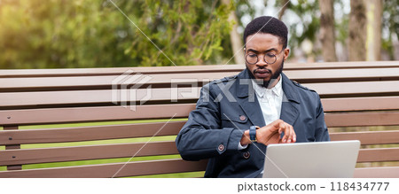 A professional black man uses his laptop on a park bench, checking time. The scene is tranquil and modern, emphasizing outdoor work-life balance. A professional black man uses his laptop on a park bench, checking time. The scene is tranquil and modern, emphasizing outdoor work-life balance. 118434777