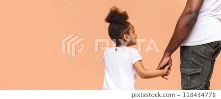 A little Black girl with a ponytail smiles as she holds her fathers hand. Her father is out of frame but his arm and hand are visible. The background is a light orange, likely studio wall, copy space 118434778