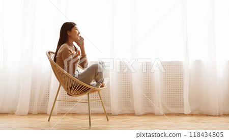 A young woman sits in a wicker chair, talking on her phone, by a window with sheer white curtains. She smiles as she talks, looking out the window. A young woman sits in a wicker chair, talking on her phone, by a window with sheer white curtains. She smiles as she talks, looking out the window. 118434805