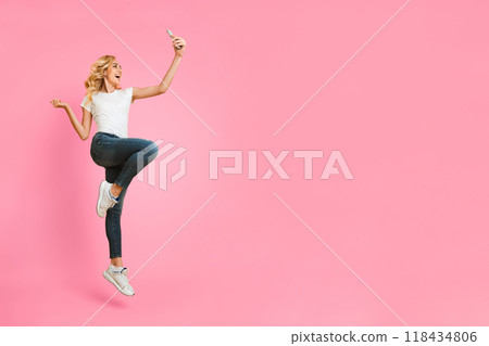 A young woman with blonde hair is jumping in the air while taking a selfie on a pink background. She is wearing a white t-shirt, blue jeans, and white sneakers. A young woman with blonde hair is jumping in the air while taking a selfie on a pink background. She is wearing a white t-shirt, blue jeans, and white sneakers. 118434806
