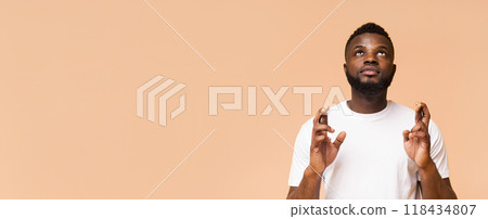 Young black man with a beard standing against a peach background. He has crossed his fingers and is looking upward, conveying a sense of hope, anticipation, or a wish being made. Young black man with a beard standing against a peach background. He has crossed his fingers and is looking upward, conveying a sense of hope, anticipation, or a wish being made. 118434807