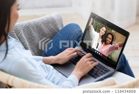 A woman is sitting on a couch at home, using a laptop to video chat with two friends. The friends are smiling and waving at the camera, while the woman is typing on the keyboard. A woman is sitting on a couch at home, using a laptop to video chat with two friends. The friends are smiling and waving at the camera, while the woman is typing on the keyboard. 118434808