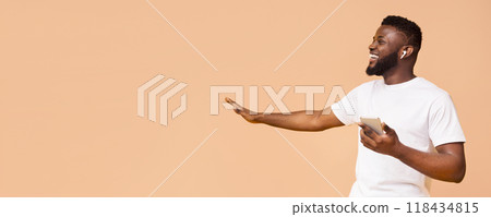 A happy young black man with a beard is wearing earbuds while holding his smartphone in one hand and gesturing with the other. He is wearing a white t-shirt and is smiling broadly, copy space 118434815