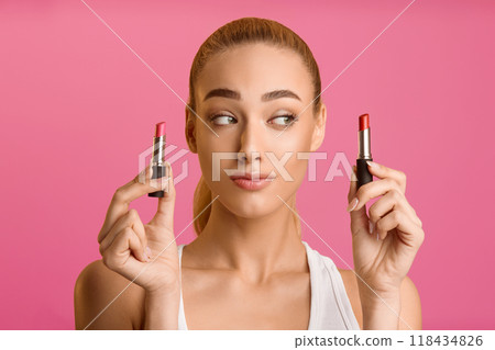 A young woman with blonde hair stands in front of a pink background. She is holding two lipsticks, one in each hand, and she is looking to the side with a thoughtful expression. A young woman with blonde hair stands in front of a pink background. She is holding two lipsticks, one in each hand, and she is looking to the side with a thoughtful expression. 118434826