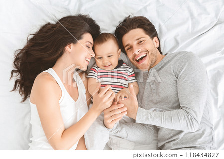 A young couple is lying on a white bed, smiling at their baby. The father is on the right side of the image and the mother is on the left. The baby is lying in the middle of the bed A young couple is lying on a white bed, smiling at their baby. The father is on the right side of the image and the mother is on the left. The baby is lying in the middle of the bed 118434827