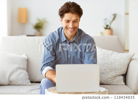 A man sits on a couch and smiles while using a laptop. A man sits on a couch and smiles while using a laptop. 118434835