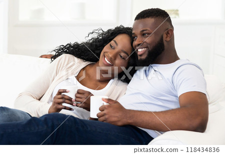 A happy couple relaxes on a white couch while holding coffee mugs, enjoying a moment of peace together. A happy couple relaxes on a white couch while holding coffee mugs, enjoying a moment of peace together. 118434836