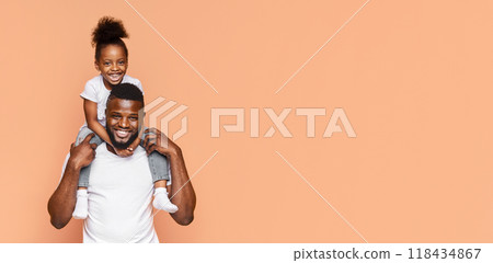 Happy Black father and his daughter posing for a photo together. The father is holding his daughter on his shoulders and they are both smiling widely. The background is a simple peach color 118434867