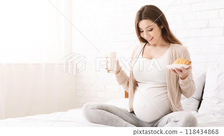 Young pregnant woman having brunch of milk and croissant, sitting on bed and looking at belly, free space Young pregnant woman having brunch of milk and croissant, sitting on bed and looking at belly, free space 118434868