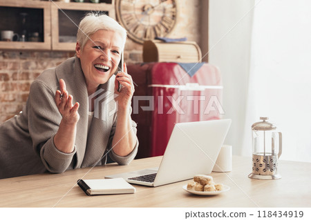 A senior woman with short grey hair is sitting at a kitchen table, smiling and talking on a cell phone. There is a laptop in front of her, along with a notebook A senior woman with short grey hair is sitting at a kitchen table, smiling and talking on a cell phone. There is a laptop in front of her, along with a notebook 118434919