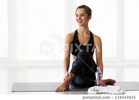A woman in activewear is sitting cross-legged on a colorful yoga mat, holding a transparent water bottle. The woman appears focused and hydrated during her yoga session. A woman in activewear is sitting cross-legged on a colorful yoga mat, holding a transparent water bottle. The woman appears focused and hydrated during her yoga session. 118434925