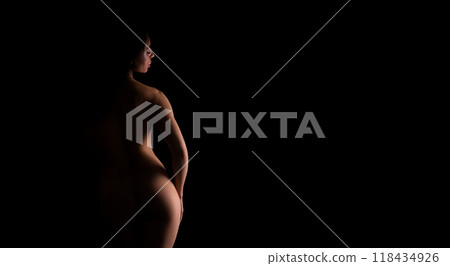 A nude woman is standing in the darkness, illuminated by a faint light source, casting shadows around her. Her figure is silhouetted against the black background. A nude woman is standing in the darkness, illuminated by a faint light source, casting shadows around her. Her figure is silhouetted against the black background. 118434926
