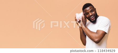 African American smiling man with a beard holding a pink piggy bank to his ear, seemingly listening to it. He is wearing a white t-shirt and has a friendly expression on his face, copy space African American smiling man with a beard holding a pink piggy bank to his ear, seemingly listening to it. He is wearing a white t-shirt and has a friendly expression on his face, copy space 118434929