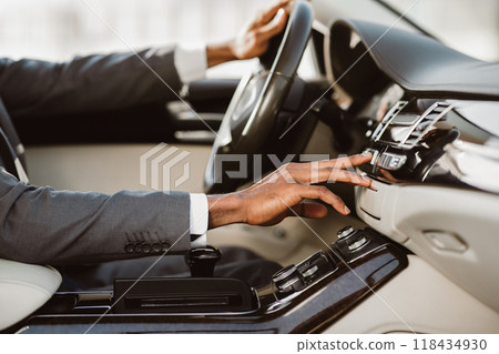 A man wearing a suit sits in the drivers seat of a car, adjusting the climate control system. His hand reaches towards the dashboard, and his other hand rests on the steering wheel, cropped A man wearing a suit sits in the drivers seat of a car, adjusting the climate control system. His hand reaches towards the dashboard, and his other hand rests on the steering wheel, cropped 118434930