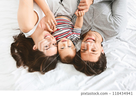 A happy family of three is lying on a bed together, holding hands. The parents are looking down at their child, who is smiling up at them. They are all wearing casual clothing, top view 118434941