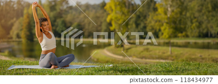 Sportswoman practicing pilates outdoors, bending in half lotus pose with arms extended, epitomizing health and flexibility, sitting on mat in green park, copy space Sportswoman practicing pilates outdoors, bending in half lotus pose with arms extended, epitomizing health and flexibility, sitting on mat in green park, copy space 118434977