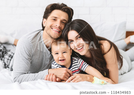 A smiling family of three lies on a bed together. The parents are both looking at the camera, while the baby is smiling at someone out of frame. The family looks relaxed and happy. A smiling family of three lies on a bed together. The parents are both looking at the camera, while the baby is smiling at someone out of frame. The family looks relaxed and happy. 118434983