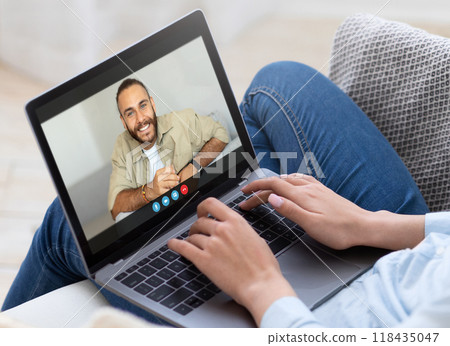 A woman is sitting on a couch and video chatting with a man on a laptop computer. She is typing on the keyboard and looking at the screen. The man is smiling and appears to be talking. 118435047