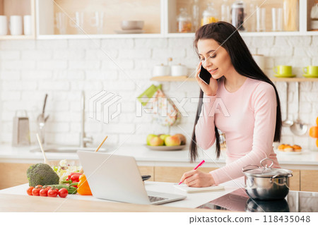 A young woman is multitasking in her modern kitchen. She is talking on the phone while taking notes, surrounded by fresh vegetables, a laptop, and a pot on the stove. 118435048