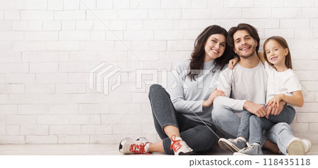 Millennial Family. Happy Young Mom, Dad And Their Cute Daughter Sitting On Floor, Posing Together Near White Brick Wall, Free Space 118435118