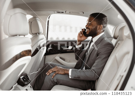 African American businessman in a suit is sitting in the backseat of a car, talking on his phone with a smile on his face. He is dressed in a gray suit, white shirt, and a dark tie 118435123