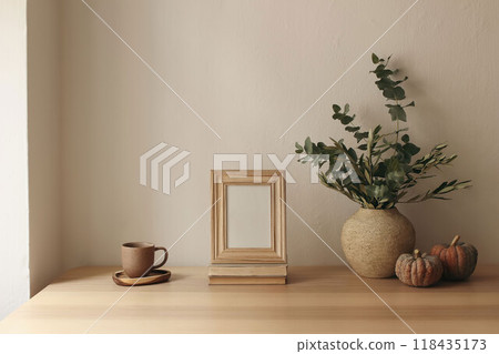 Autumn interior still life. Vase with olive eucalyptus tree branches, pumpkins. Blank wooden photo frame mockup on table. Thanksgiving decor. Scandi interior. Cup of tea, coffee. Beige wall background 118435173