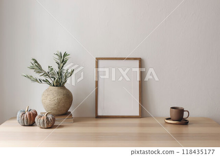 Elegant autumn interior still life. Vase with olive branches, pumpkins. Blank vertical wooden picture frame mockup on table. Home office. Scandinavian interior. Cup of tea, coffee. White wall 118435177