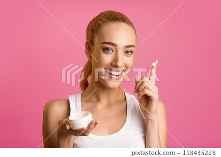 A young woman, with blonde hair pulled back, smiles at the camera as she applies a white cream to her finger. She is holding an empty container in her other hand. The background is a vibrant pink. A young woman, with blonde hair pulled back, smiles at the camera as she applies a white cream to her finger. She is holding an empty container in her other hand. The background is a vibrant pink. 118435228