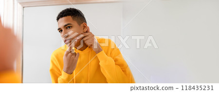 A young African American man wearing a yellow hoodie is in a bathroom, focusing on his skincare routine by inspecting his skin in the mirror and gently touching his face. A young African American man wearing a yellow hoodie is in a bathroom, focusing on his skincare routine by inspecting his skin in the mirror and gently touching his face. 118435231