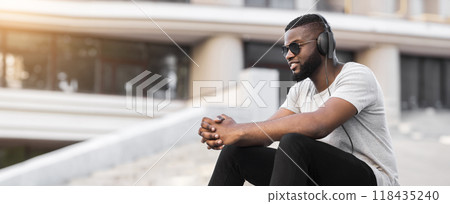 A young man wearing headphones sits on a step in an urban environment. He is wearing sunglasses and a white shirt and is looking to the side. 118435240