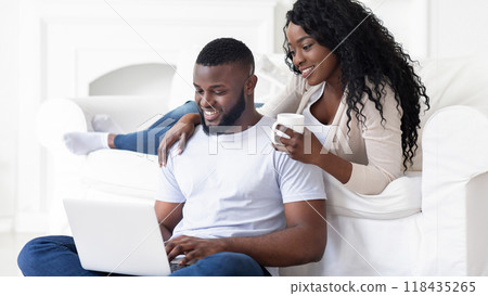 A couple relaxes on a white couch, one working on a laptop while the other holds a mug of coffee. 118435265