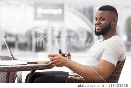 A man sits at a table outside a cafe, smiling as he looks to the side and uses his phone. A man sits at a table outside a cafe, smiling as he looks to the side and uses his phone. 118435266