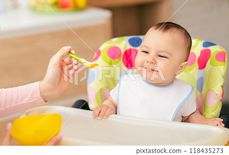 A mom is feeding her baby boy who is sitting in a high chair in the kitchen. The scene is warm and homey, emphasizing the nurturing bond during mealtime. A mom is feeding her baby boy who is sitting in a high chair in the kitchen. The scene is warm and homey, emphasizing the nurturing bond during mealtime. 118435273
