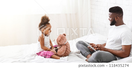 Black father and his little daughter are sitting on a bed, both smiling. The daughter is holding a stuffed bear, while the father is looking at his daughter with a tablet in his lap 118435274