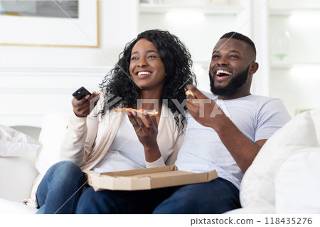 A couple relaxes on a couch, laughing and enjoying pizza while watching television together at home. 118435276