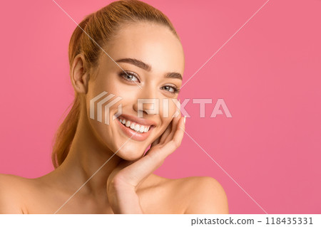 The image shows a close-up portrait of a young woman with long brown hair pulled back in a ponytail. She is smiling brightly and has her hand resting on her cheek. The background is solid pink The image shows a close-up portrait of a young woman with long brown hair pulled back in a ponytail. She is smiling brightly and has her hand resting on her cheek. The background is solid pink 118435331