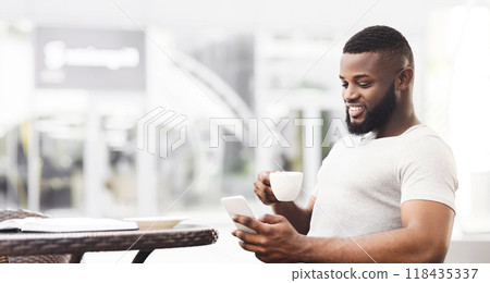 A man with a beard sits at a cafe table, smiling while looking at his smartphone and holding a cup of coffee. A man with a beard sits at a cafe table, smiling while looking at his smartphone and holding a cup of coffee. 118435337