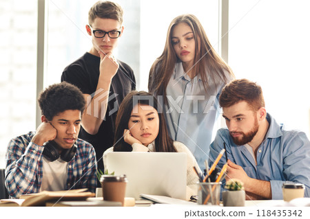 A group of five young professionals is gathered around a laptop, collaborating intently on a project. Some appearing pensive and others focused, they work together in a sunlit office setting A group of five young professionals is gathered around a laptop, collaborating intently on a project. Some appearing pensive and others focused, they work together in a sunlit office setting 118435342