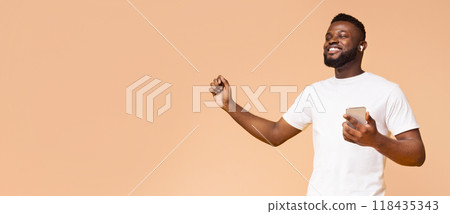 African American young man in a white t-shirt dances and smiles while holding a smartphone and wearing earbuds. He enjoying music in front of a light beige background, copy space 118435343
