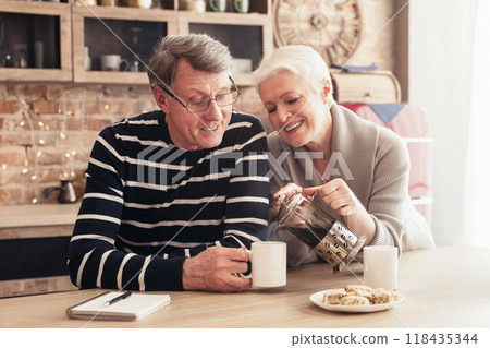 Sincere love. Happy senior couple sitting at table, drinking tea in kitchen, empty space, sun flare 118435344