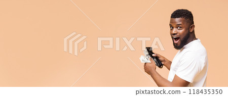 African American young man with a beard is seen holding a wallet and a wad of cash while looking towards the camera with a surprised expression, copy space 118435350