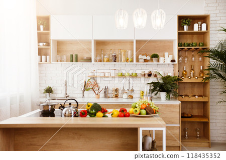 A modern kitchen island with fresh fruit, vegetables on display. The island is made of light wood and has a white countertop. There is kettle on the stovetop, other kitchen supplies in the background. A modern kitchen island with fresh fruit, vegetables on display. The island is made of light wood and has a white countertop. There is kettle on the stovetop, other kitchen supplies in the background. 118435352