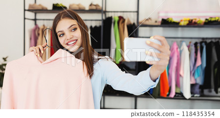 A cheerful young woman is holding up a pink sweater and using her smartphone to take a selfie in a modern clothing store, surrounded by various colorful garments hanging in the background. 118435354